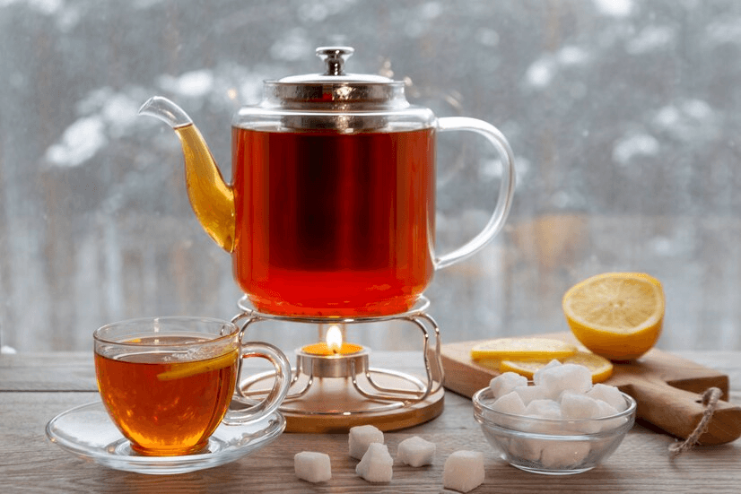 A warm glass teapot filled with vibrant English Breakfast tea on a tea warmer, accompanied by a glass teacup with a slice of lemon, sugar cubes, and a halved lemon on a wooden cutting board, placed by a snowy window for a cozy winter ambiance.